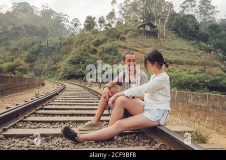Side view of couple on vacation sitting on rails smiling and looking at each other near tropical green plants on hills in Sri Lanka Asia Stock Photo