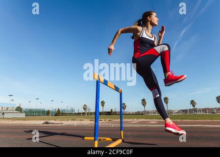 Side view of strong young Woman in sportswear leaping over hurdle against blue sky during workout on stadium Stock Photo