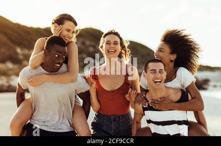Happy friends standing together enjoying vacation. Multiethnic group of friends having fun piggy riding standing outdoors. Stock Photo