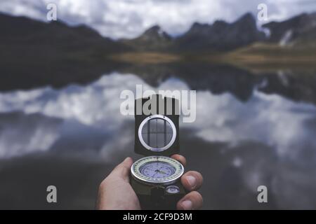 Hand of anonymous traveler holding compass against tranquil mountain lake on cloudy day in Spanish countryside Stock Photo