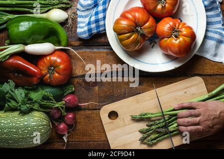 Top view of crop person cutting green asparagus on cutting board on plank wooden table with ripe red tomatoes placed on white plate and mixed colorful vegetables placed aside Stock Photo