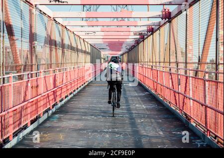 Back view of anonymous cyclist with helmet riding bike among red metal bridge structure in New York city Stock Photo