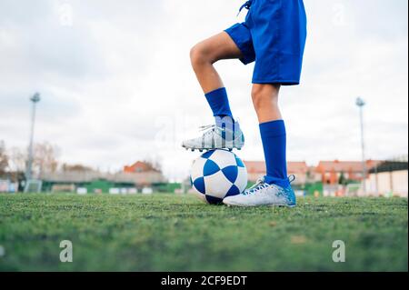 Low angle side view of unrecognizable teenage girl in white and blue uniform and goalkeeper gloves kicking ball while training alone in football arena at stadium in daytime Stock Photo