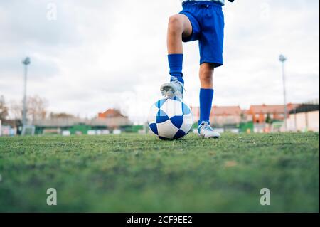Low angle side view of unrecognizable teenage girl in white and blue uniform and goalkeeper gloves kicking ball while training alone in football arena at stadium in daytime Stock Photo
