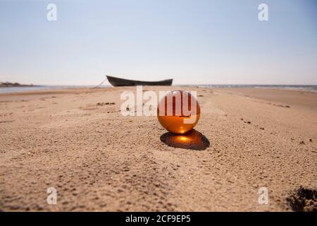 Orange glass ball on sand near the sea. Abstract conceptual photo. Stock Photo