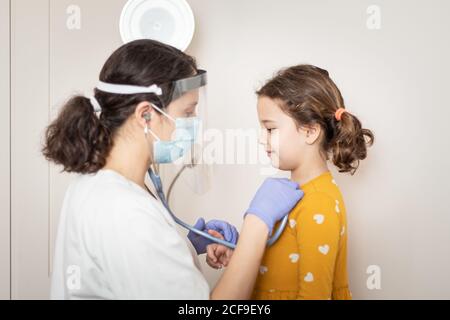 Side view of female doctor in white uniform and in latex gloves and protective mask examining with stethoscope lungs of small girl Stock Photo