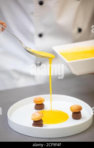 Crop cooker in uniform holding bowl and pouring yellow syrup from spoon into white plate with chocolate pastries in form of mushrooms Stock Photo