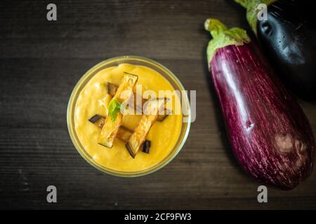 Top view of cup of tasty puree with slices of roasted eggplant and herb placed on lumber table near raw vegetables in kitchen Stock Photo