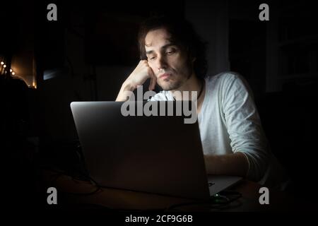 Concentrated young male remote specialist in casual wear talking on mobile phone and using laptop while working on project in dark room at home during evening time Stock Photo