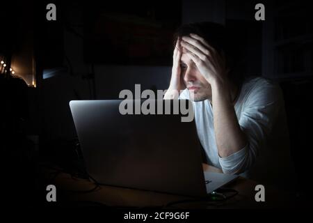 Exhausted young male in casual wear sitting at table with laptop while working late on remote project in dark room at home Stock Photo