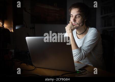 Concentrated young male remote specialist in casual wear talking on mobile phone and using laptop while working on project in dark room at home during evening time Stock Photo