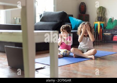 High angle of positive little sisters sitting in lotus pose with outstretched arms while watching video tutorial and practicing yoga together in living room at home Stock Photo