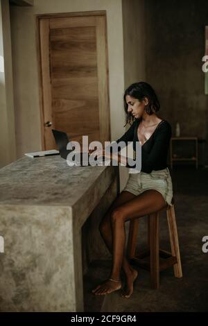 Serious focused youthful ethnic female typing on laptop while sitting at table on table kitchen modern marble countertop Stock Photo