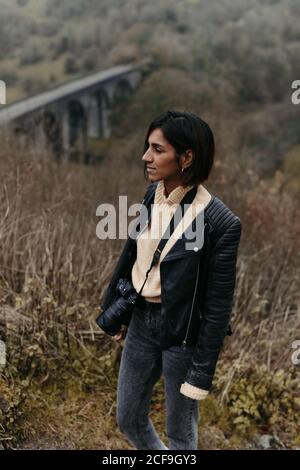 Thoughtful touristic Woman with camera in stylish black leather jacket walking in valley with dry grass and old bridge in United Kingdom Stock Photo