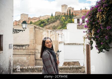 Side view of pleased Asian female tourist in stylish coat looking at camera with ancient rocked buildings and beautiful castle on background at Alcazar of Alhambra in Granada Stock Photo
