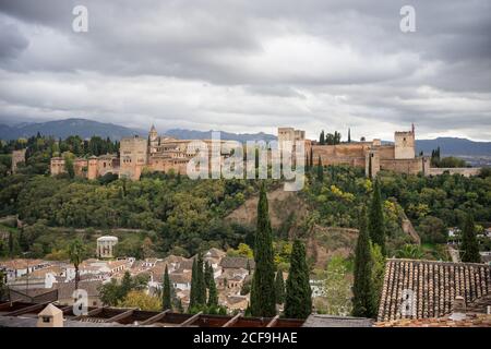 Powerful ancient rocked castle Alcazaba of Alhambra on big overgrown of green trees near old town with mountains and cloudy sky on background at Granada Stock Photo