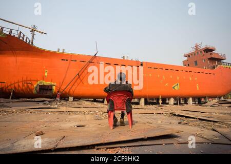 Bangladesh - January, 20 2019: Back view of anonymous ethnic man sitting on chair and talking on smartphone while supervising shipbuilding process against cloudless sky in harbor Stock Photo