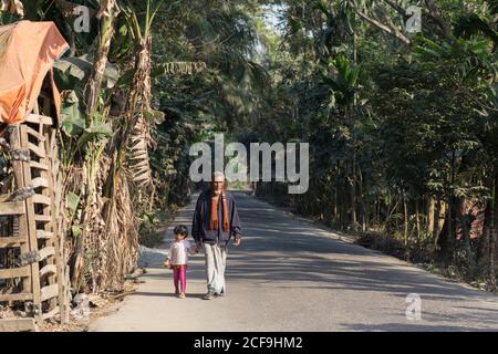 Serious ethnic aged ethnic man and kid holding hands and walking on asphalt road through tropical forest in Barisal in Bangladesh Stock Photo