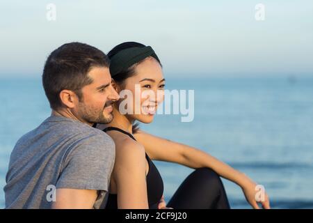 Side view of cheerful young multiracial couple in sportswear sitting on sandy beach while resting after training and enjoying time together Stock Photo
