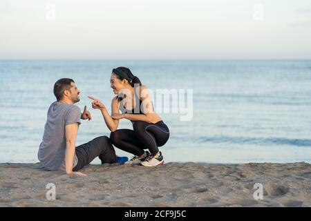 Side view of cheerful young multiracial couple in sportswear sitting on sandy beach while resting after training and enjoying time together Stock Photo