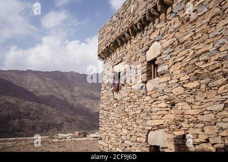 Woman leaning out of a window of ancient stone wall of historic building of Marble Village in Al Bahah against background of rocky terrain and cloudy sky in summer in Saudi Arabia Stock Photo