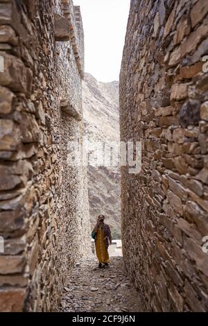 Distant Woman in colorful clothes walking between shabby ancient walls of historic buildings of Marble Village in Al Bahah locating on mountain terrain in Saudi Arabia Stock Photo