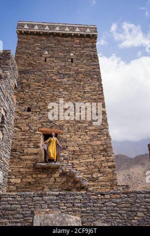 From below of monumental ancient building with remote female tourist coming out of doorway in yellow dress while enjoying hot sunny day in Marble Village in Al Bahah Stock Photo