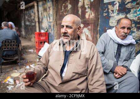 Cairo, Egypt - December 14, 2019: Old man holding glass of drink chatting with friends relaxing in street Stock Photo