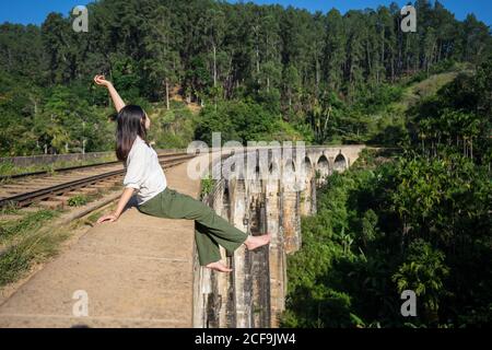 Side view of active pleasant Asian touristic Woman sitting on ancient bridge dangling leg and raising hand up along old railway in green place in Nine Arches Bridge, Ella, Sri Lanka Stock Photo