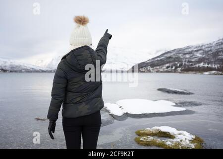 Back view of anonymous Woman in warm down jacket and hat pointing with finger while standing on beach against white and brown snowy hills and tranquil cool water of Kaldfjorden lake in Norway Stock Photo