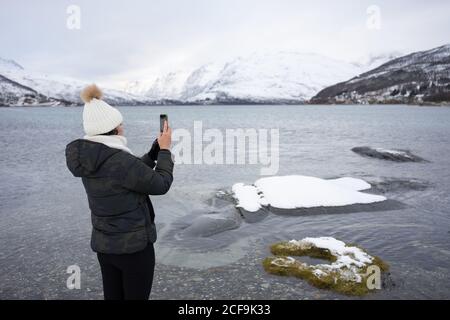 High angle back view of unrecognizable female in warm clothes taking shot with smartphone of amazing wintry scenery of highland while standing on shore of Kaldfjorden lake in Norway Stock Photo
