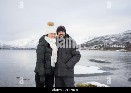 Joyful couple in warm down jacket and hats hugging and smiling at camera while standing on beach against white and brown snowy hills and tranquil cols water of Kaldfjorden lake in Norway Stock Photo