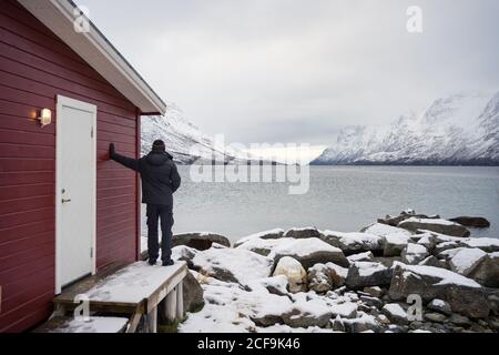 From behind anonymous man in warm black and white clothes standing on stairs beside small red country house and contemplating beautiful wintry landscape of Ersfjordbotn lake in Norway Stock Photo