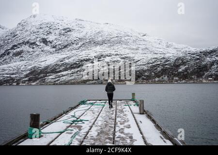 Back view of anonymous Woman in warm clothes contemplating while standing on edge of pier against small red country houses on wintry Ersfjordbotn lake in Norway Stock Photo