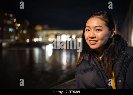 Delighted Asian Woman smiling and looking at camera while standing on dark embankment on blurred background of river and Ponte Vecchio at night in Florence, Italy Stock Photo