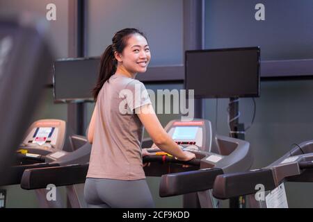 Back view of slim happy young ethnic sportsWoman in active wear looking at camera while running on treadmill while exercising in spacious modern fitness center Stock Photo