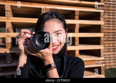 Cheerful young Asian female photographer in casual wear and wristwatch taking photo on digital photo camera while standing near wooden fence and looking at camera Stock Photo