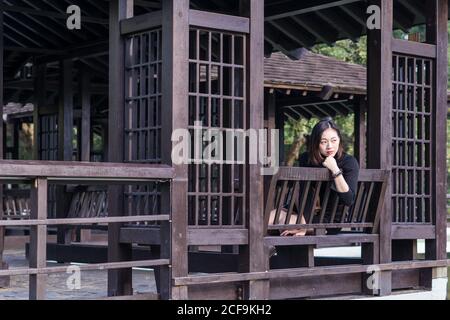 Dreamy young Asian female sitting on wooden bench on fenced terrace near trees leaned on hand and looking away in daylight Stock Photo