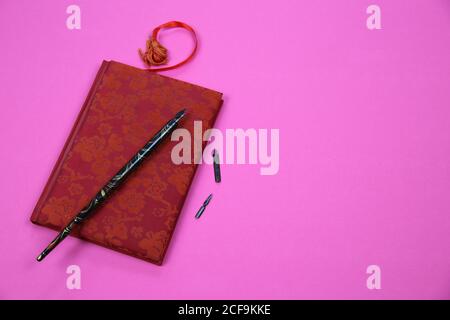 View on isolated notebook with red floral pattern cover, old feather ink pen and bookmark on pink background with copy space for text Stock Photo