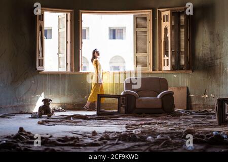 Side view of young ethnic female in yellow dress standing near open window in messy room of old uninhabited house with remains of furniture and garbage on floor Stock Photo