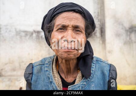 Maubisse, East Timor - AUGUST 10, 2018: Poor old Asian female in black headscarf sitting against concrete wall and looking at camera Stock Photo