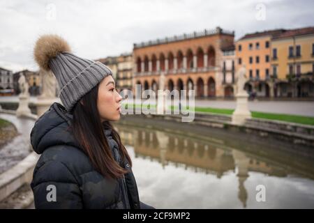 Side view of young Asian resting lady in warm clothing and hat walking at beautiful Prato della Valle park at Padova at Italy