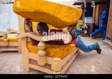 Ruanda, African - December 14, 2019: African American guy carrying furniture lying on sofa holding pillow on market stall Stock Photo