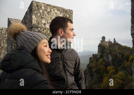 Thoughtful man and smiling Asian Woman in knitted hat with pompom looking away and enjoying amazing landscape of ancient castle in San Marino, Italy Stock Photo