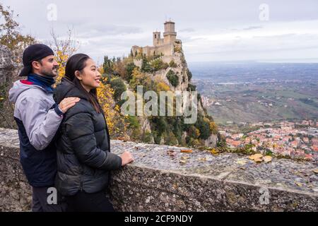 Amorous man embracing smiling Asian Woman in knitted hat with pompom leaning on stony fence on hill with amazing landscape of ancient castle in San Marino, Italy Stock Photo