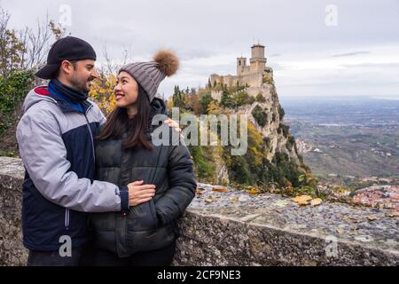 Amorous man embracing smiling Asian Woman in knitted hat with pompom leaning on stony fence on hill with amazing landscape of ancient castle in San Marino, Italy Stock Photo