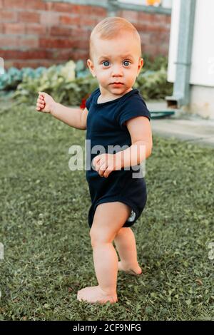 Adorable baby boy in dark blue baby suit looks at camera, while trying to keep his balance Stock Photo