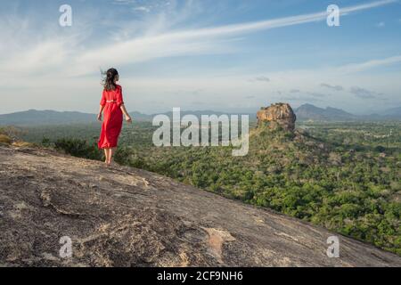 Back view of unrecognizable Woman in red dress enjoying amazing view of lonely rock fortress Sigiriya in middle of plateau with dense green tropical forest from mountain Pidurangala under blue sky in Sri Lanka Stock Photo