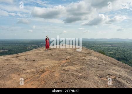 Woman in red dress raising arms up enjoying amazing view of lonely rock fortress Sigiriya in middle of plateau with dense green tropical forest from mountain Pidurangala under blue sky in Sri Lanka Stock Photo