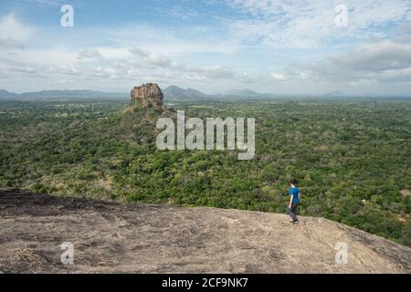 Side view of unrecognizable man enjoying amazing view of lonely rock fortress Sigiriya in middle of plateau with dense green tropical forest from mountain Pidurangala under blue sky in Sri Lanka Stock Photo